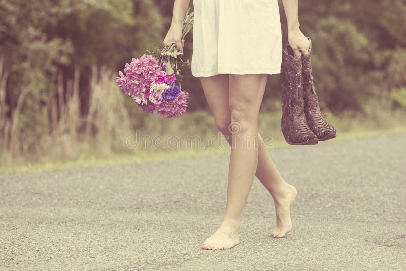 Una bella donna che indossa un abito bianco, cammina lungo la strada portando i suoi stivali e i fiori da un triste rottura.