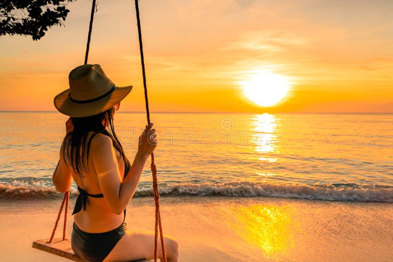 Sexy woman wear bikini and straw hat swing the swings at tropical beach on summer vacation at sunset. Girl in swimwear sit on swings and watching beautiful sunset. Summer vibes. Woman travel alone