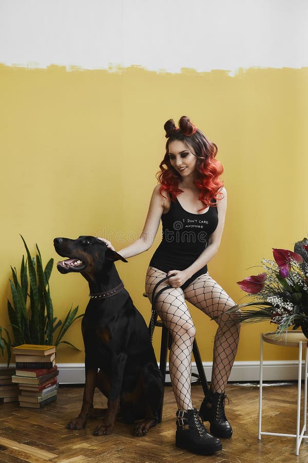 Sexy young model woman with red hair and with perfect body in black stylish bodysuits and in mesh pantyhose sits on the chair near big Doberman.