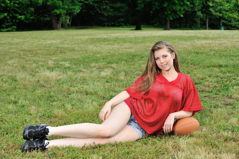 young Caucasian woman in red football jersey, and denim jeans, resting on an American football in a field of grass - smiling. young Caucasian woman in red football jersey, and denim jeans, resting on an American football in a field of grass - smiling