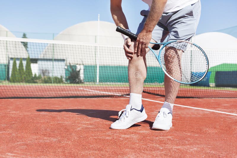 Sezione bassa dell'uomo maturo che tiene la racchetta di tennis mentre soffrendo dal dolore del ginocchio sul campo da tennis ros