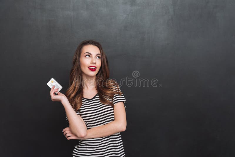 Señora joven de pensamiento que se coloca sobre la pared gris que sostiene la tarjeta de débito fotos de archivo Señora joven de pensamiento que se coloca sobre la pared gris que sostiene la tarjeta de débito fotos de archivo