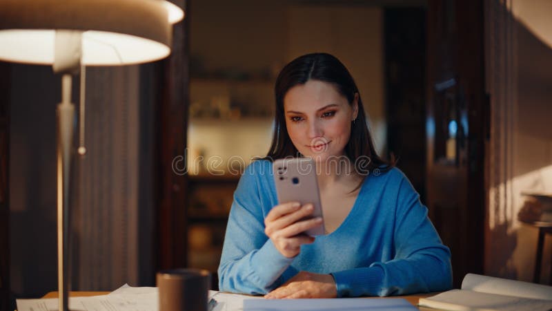 Señora leyendo mensajes de teléfono celular sentada en un lugar de trabajo oscuro. Mujer sonriente fotos de archivo libres de regalías