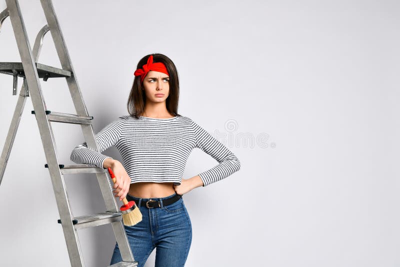 Sfrustowana Młoda brunetki dziewczyna z muśnięciem i stepladder - ile pracy mu robić na malować ściany fotografia stock Sfrustowana Młoda brunetki dziewczyna z muśnięciem i stepladder - ile pracy mu robić na malować ściany fotografia stock