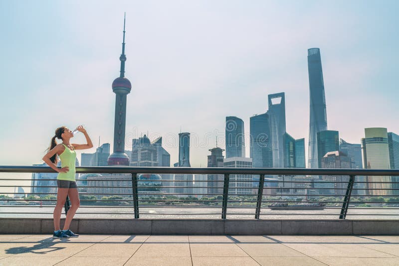 Shanghai skyline fitness woman thirsty drinking water bottle on sunny day, China. Asian jogger thirst quenching drink