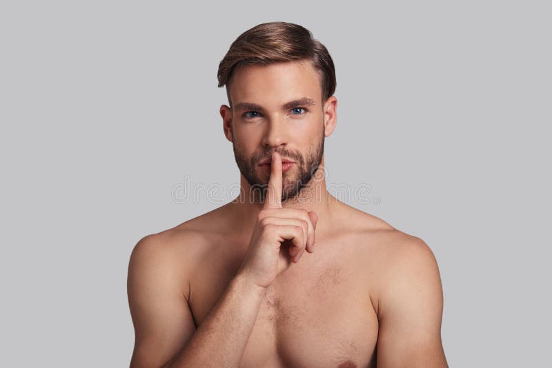 Thoughtful young man looking at camera and keeping index finger by his lips while standing against grey background. Thoughtful young man looking at camera and keeping index finger by his lips while standing against grey background