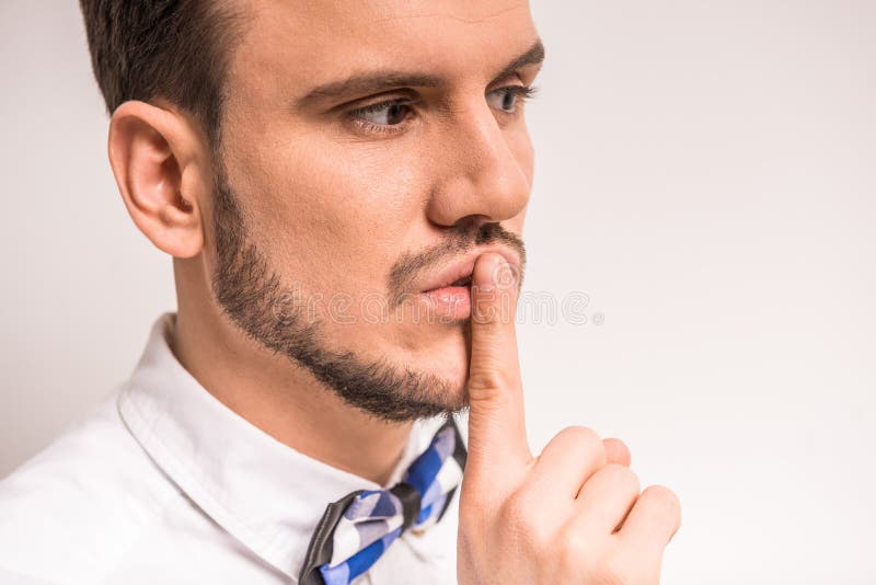 Keep silence! Handsome young man in white shirt is holding finger on lips while standing against grey background. Keep silence! Handsome young man in white shirt is holding finger on lips while standing against grey background.