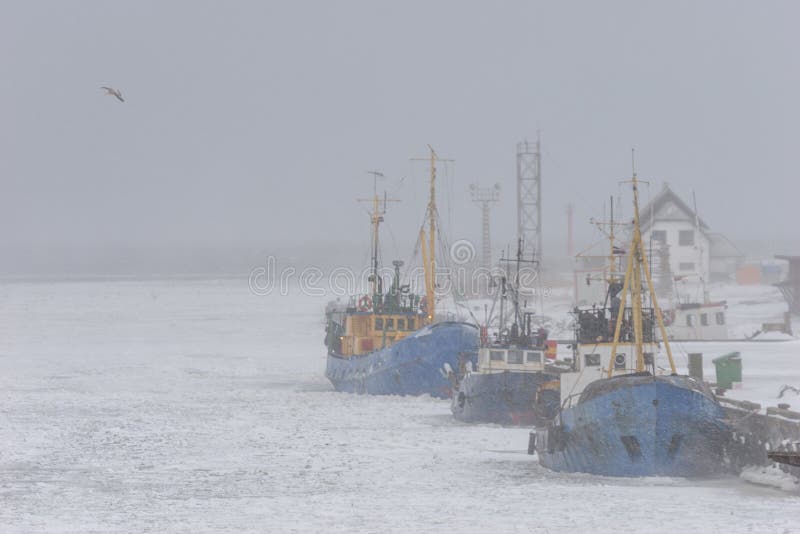 Ships of fishermen in the port on a frozen river in winter through a strong blizzard to the horizon. Ships of fishermen in the port on a frozen river in winter through a strong blizzard to the horizon