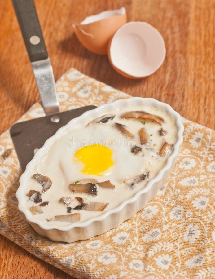 Top view, close distance of a hot, freshly prepared, homemade, shirred egg, mushroom-cream Brûlée in a white ramekin, on a spatula  over printed napkin on a wood cutting board with one egg shell. Top view, close distance of a hot, freshly prepared, homemade, shirred egg, mushroom-cream Brûlée in a white ramekin, on a spatula  over printed napkin on a wood cutting board with one egg shell