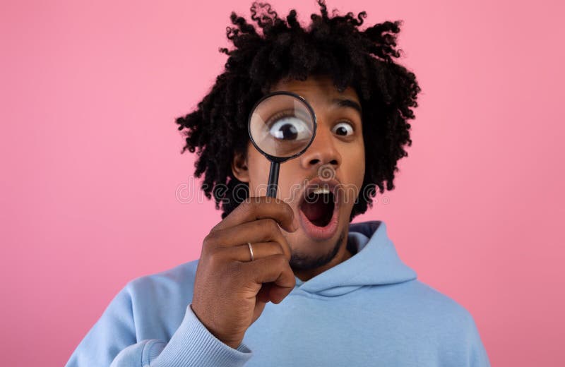 Shocked black teen guy looking through magnifying glass on pink studio background. Secret, doubt concept