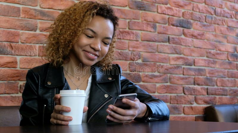 A young modern beautiful African-American girl is smiling talking on the phone and drinking a drink from a white cup