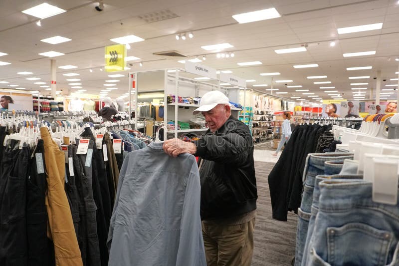 Mt. Kisco, Westchester County, New York. 4 November 2023. Senior guy with white ball cap and black winter jacket holds pale dusty blue collar shirt by hangar. Pants and shirts with sale tags stuffed on racks around him in bright big box store. Mt. Kisco, Westchester County, New York. 4 November 2023. Senior guy with white ball cap and black winter jacket holds pale dusty blue collar shirt by hangar. Pants and shirts with sale tags stuffed on racks around him in bright big box store.