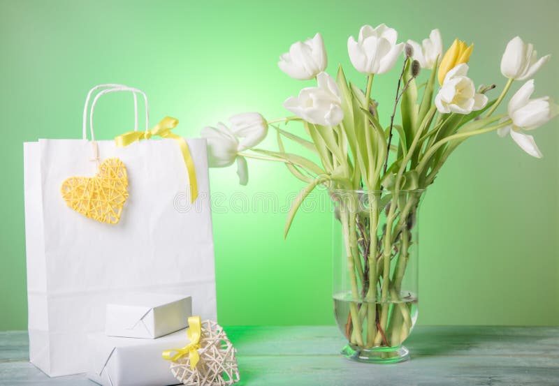 Shopping bag, yellow heart and ribbon, gift and white bouquet tulips in a vase on green background.