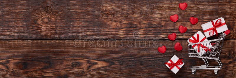 Shopping trolley, gifts, hearts on brown wooden background. 14 February. Valentine`s Day. Extra wide panorama banner background