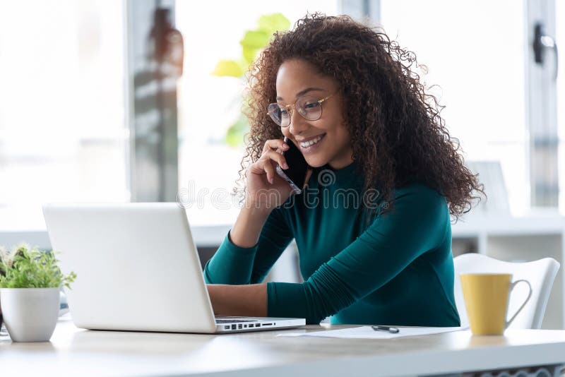 Beautiful young business woman talking on mobile phone while working with her laptop in the office