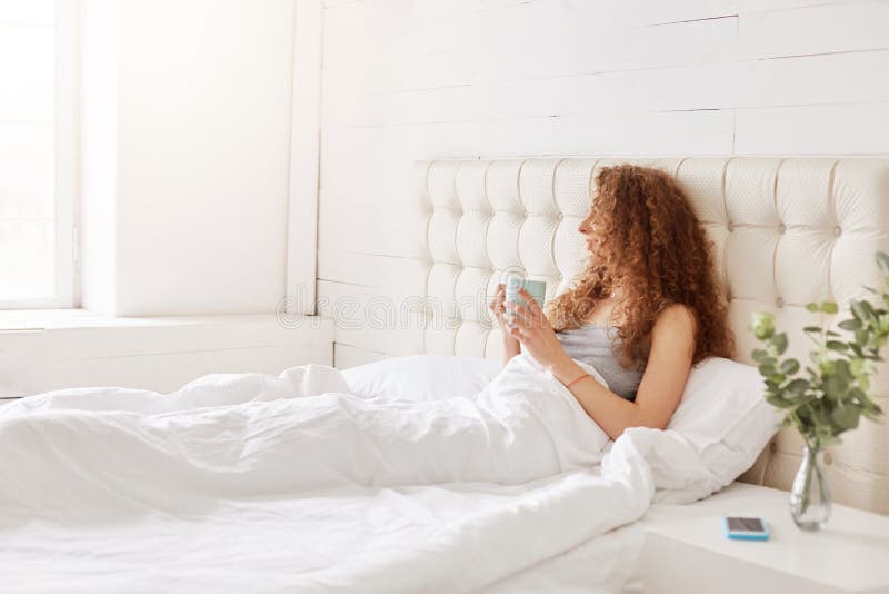 Shot of carefree restful female sits in comfortable bed with cup of hot coffee or tea, sits in spacious white bedroom, enjoys good morning. People, lifestyle, rest and starting new working day concept