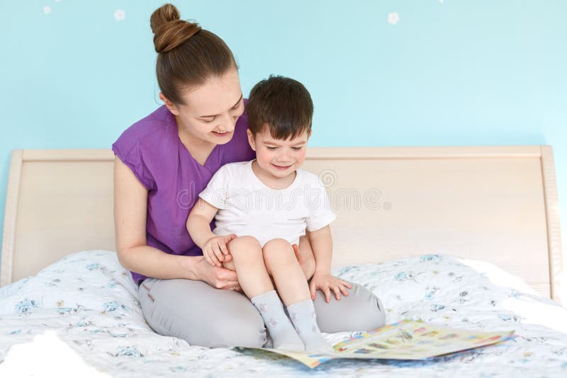 Shot of caring mother on maternity leave and small boy read fairy tale before sleep, pose at bed, view photos, enjoy domestic