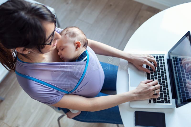 Pretty young mother with her baby in sling working with laptop at home