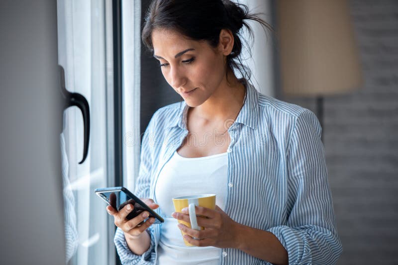 Pretty young woman using her mobile phone while drinking coffee near to the window in the living room at home