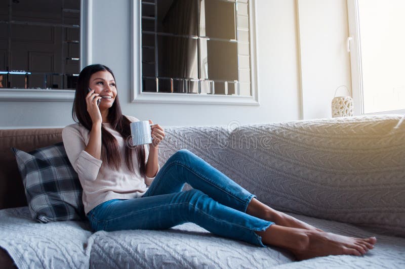 Have you heard latest news? Full length of beautiful young woman holding coffee mug and talking on mobile phone with smile while sitting on the sofa in home interior. Have you heard latest news? Full length of beautiful young woman holding coffee mug and talking on mobile phone with smile while sitting on the sofa in home interior