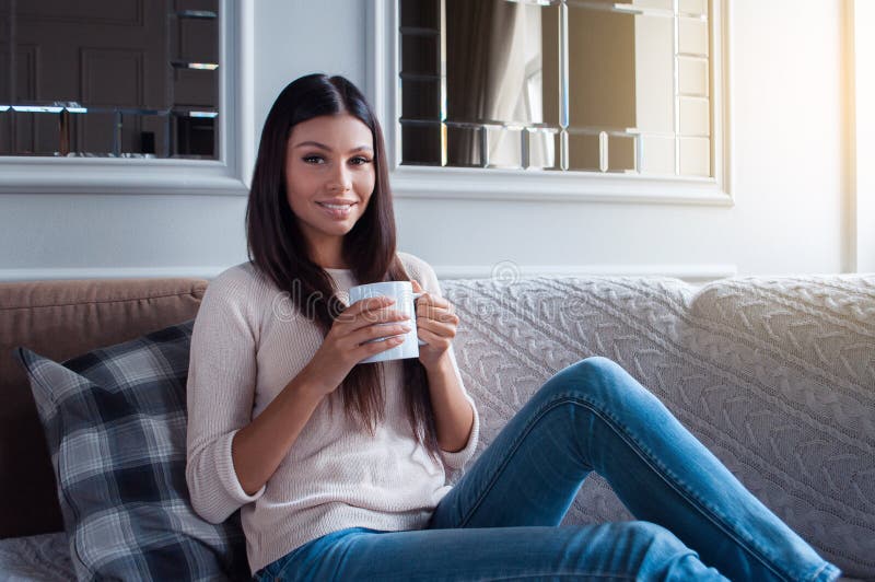 Spending perfect morning at home. Beautiful young woman holding coffee mug and looking at camera with smile while sitting on the sofa in home interior. Spending perfect morning at home. Beautiful young woman holding coffee mug and looking at camera with smile while sitting on the sofa in home interior