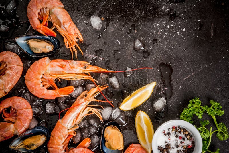 Prepared for cooking dinner seafood - shrimp and mussels on ice, on black concrete table, with lemon and seasonings. Top view, copy space