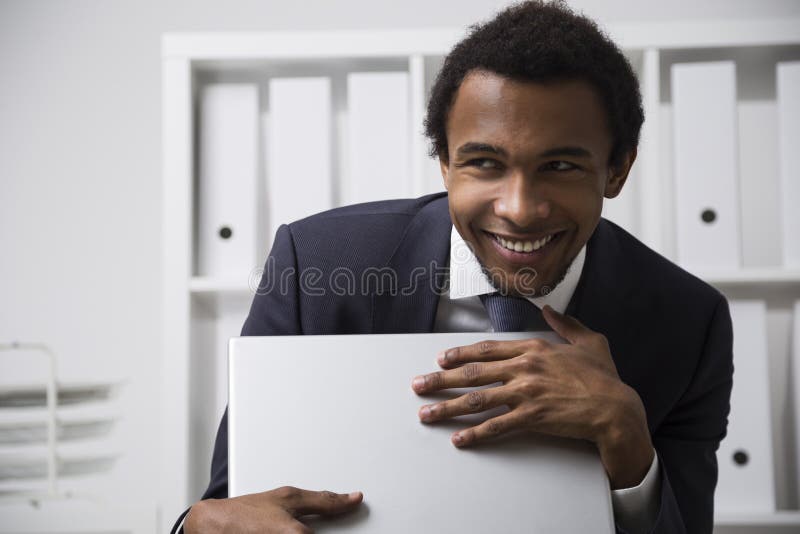 Shy and smiling African American clerk. Portrait of a very shy African American office clerk working in a white office, smiling and grasping his laptop as a stock images Shy and smiling African American clerk. Portrait of a very shy African American office clerk working in a white office, smiling and grasping his laptop as a stock images