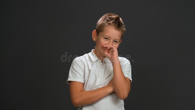 Shyly laughing boy covers his face with his hand looking at the camera wearing white polo shirt and black pants isolated