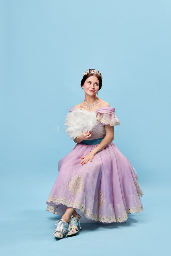 Shyness. Young elegant girl in lilac color medieval dress as young queen or princess on blue background. Eras comparison stock photography