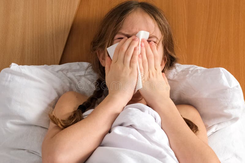 Sick little girl sneezing and blowing her nose, lying on bed at home, close up royalty free stock image