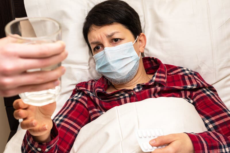 Sick woman in medical mask lying on the bed is given glass of water to take pills. Family care for the sick concept stock photo Sick woman in medical mask lying on the bed is given glass of water to take pills. Family care for the sick concept stock photo