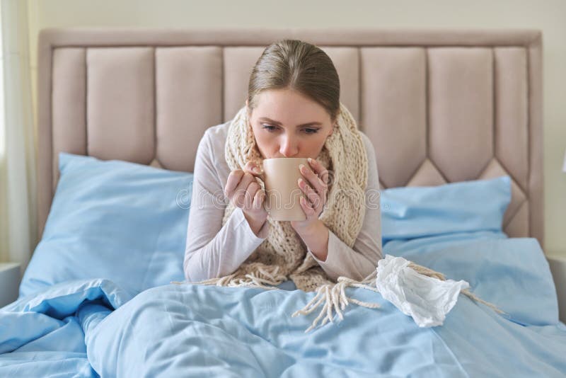 Sick young woman sitting at home in bed with hot cup of tea and handkerchief