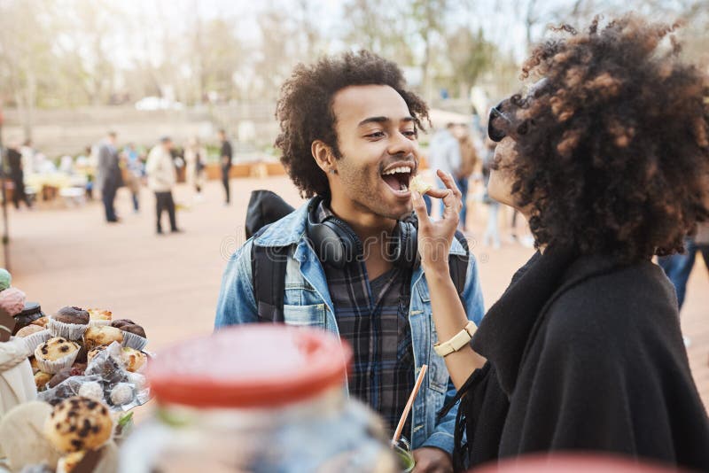 Side-view of cute african-american couple in love having fun in park during food festival, standing near counter and picking something to eat. Girlfriend feeds guy with sweet candy. Side-view of cute african-american couple in love having fun in park during food festival, standing near counter and picking something to eat. Girlfriend feeds guy with sweet candy.