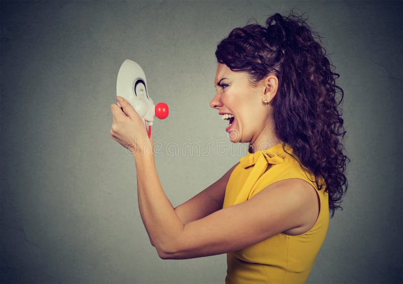 Side profile of an angry young woman screaming at happy clown mask isolated on gray background. Side profile of an angry young woman screaming at happy clown mask isolated on gray background