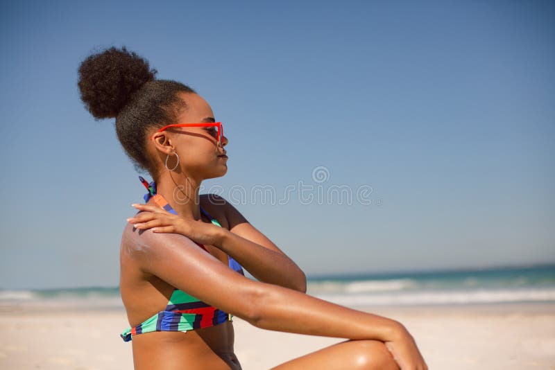 Side view of African american woman in bikini applying sunscreen lotion on shoulder at beach in the sunshine