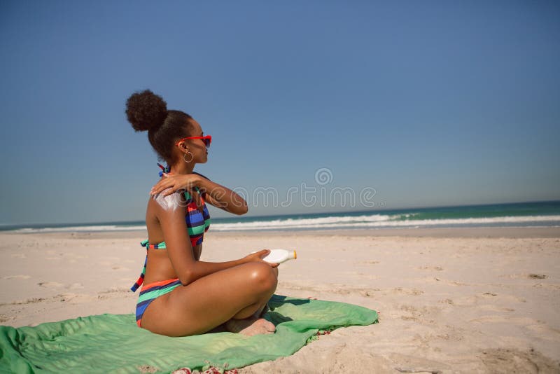 Side view of African american woman in bikini applying sunscreen lotion on shoulder at beach in the sunshine