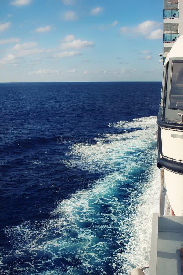 Side view from balcony deck of big cruise ship sailing ocean or sea and leaving water swirl and churn trails with blue sky on