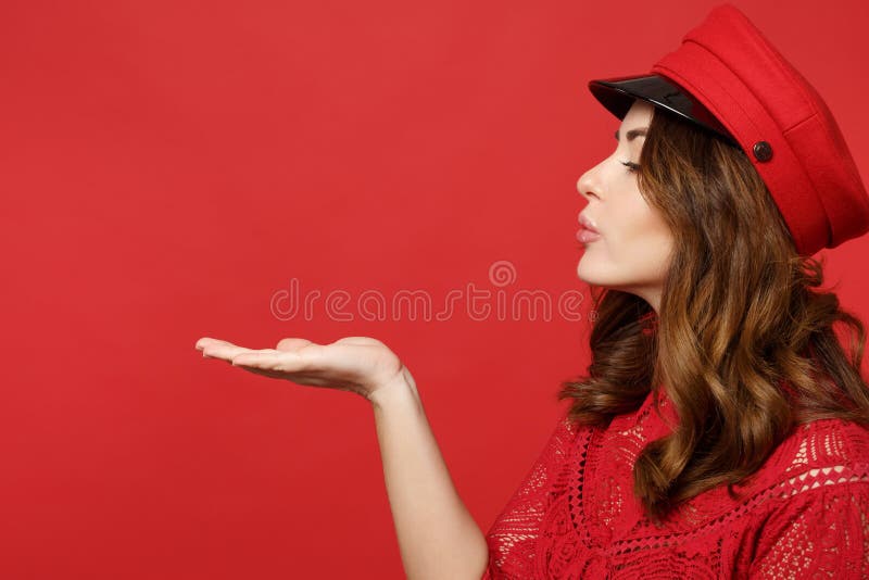 Side view of beautiful cute young woman in lace dress, cap blowing sending air kiss isolated on bright red wall stock image
