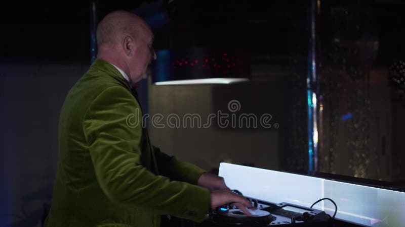 Side view of cheerful old Caucasian man mixing music on controller and waving to visitors in night club. Senior male DJ