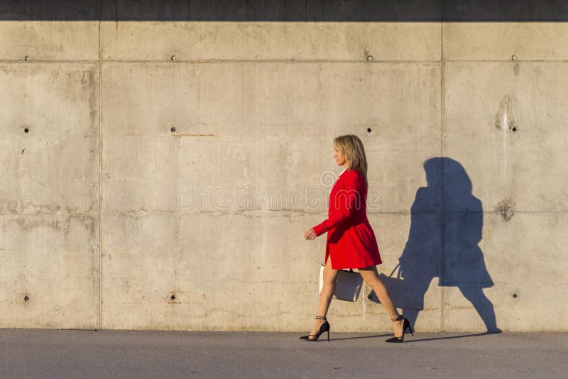 Side view of an elegant woman wearing red jacket, skirt and holding a white handbag while walking in the street in a sunny day