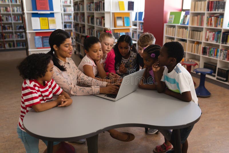 Female teacher teaching schoolkids on laptop at table in school library stock photography Female teacher teaching schoolkids on laptop at table in school library stock photography