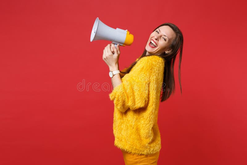 Side view of laughing young woman in yellow fur sweater holding megaphone in hands isolated on bright red wall stock photography