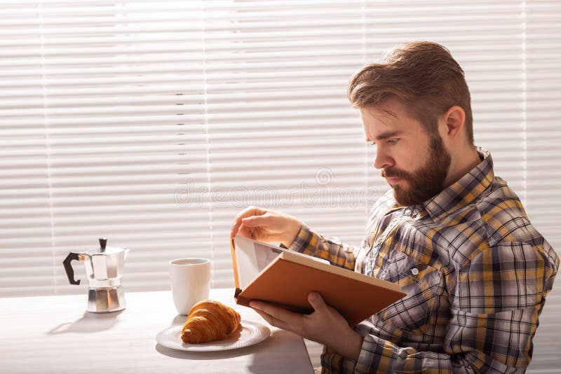 Side view of pensive young bearded hipster man reading book and having dinner with croissant and cup of coffee on