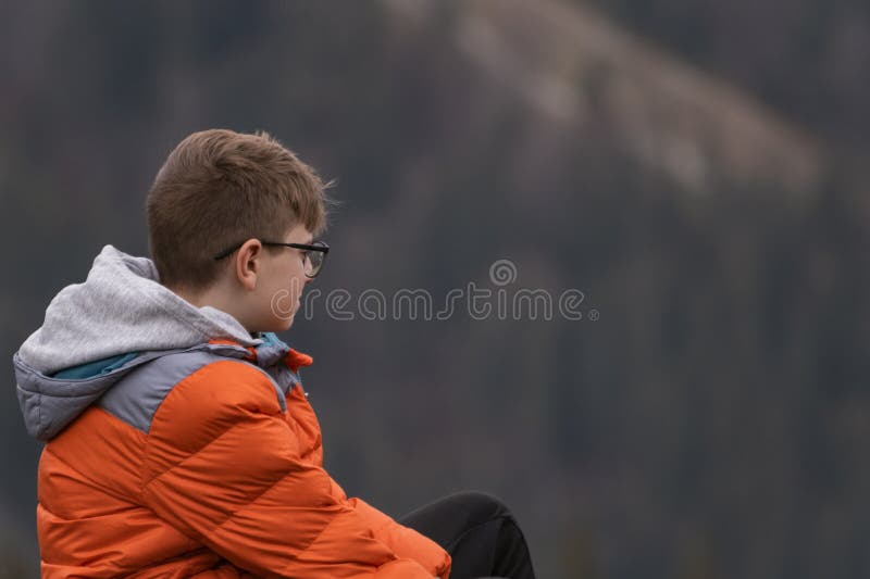Side view portrait of boy wears sporty orange jacket and glasses. Guy with brown hair outdoors. Mountains view
