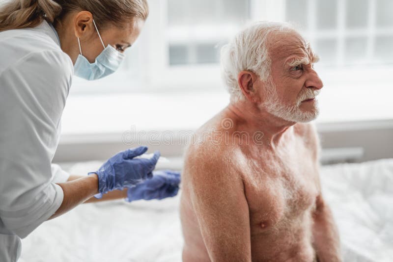 Shirtless old man receiving injection in back while sitting on hospital bed...