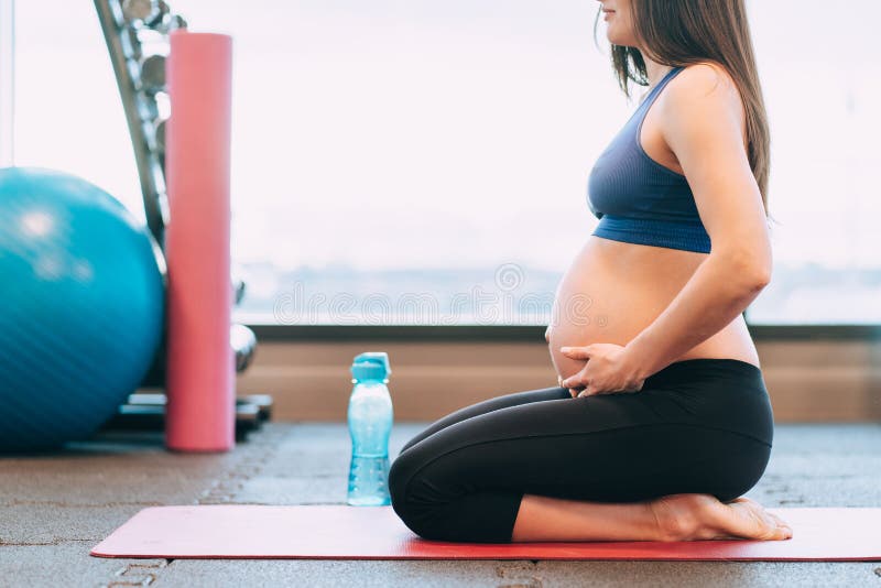 Young pregnant woman in sportswear meditate sitting holding belly on pink mat after practising yoga near bottle at gym.