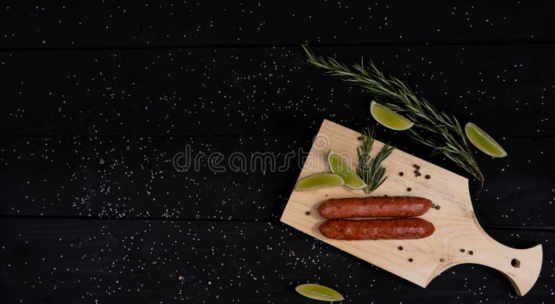 Side view of a sausage with rosemary leaves and lemon peels on a wooden board, black background, copy space