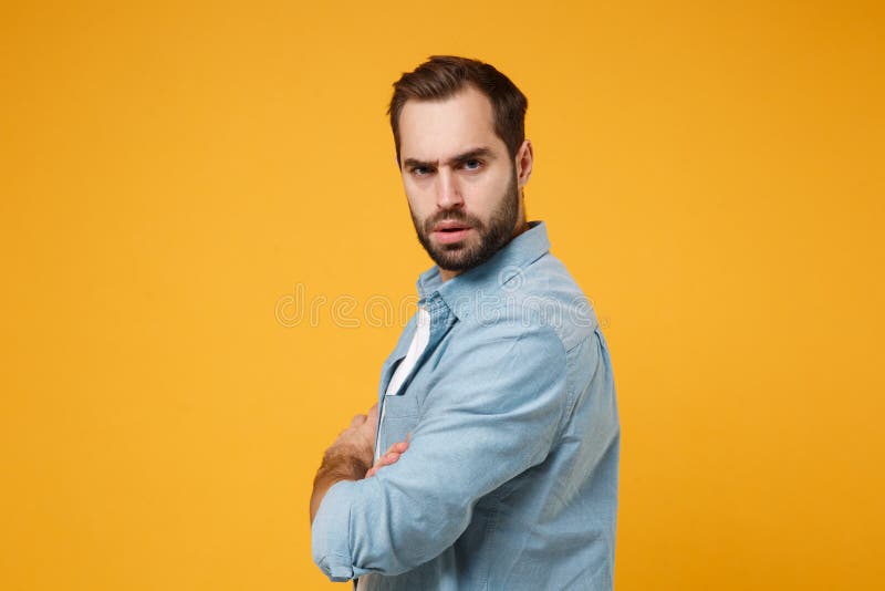 Side view of serious young bearded man in casual blue shirt posing isolated on yellow orange background in studio