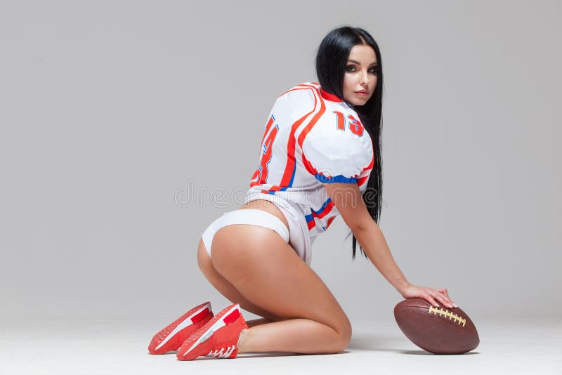 Sexy sporty brunette dressed in bikini and American football uniforms and jersey T-shirt sitting on the floor with a ball grey background. Sexy sporty brunette dressed in bikini and American football uniforms and jersey T-shirt sitting on the floor with a ball grey background