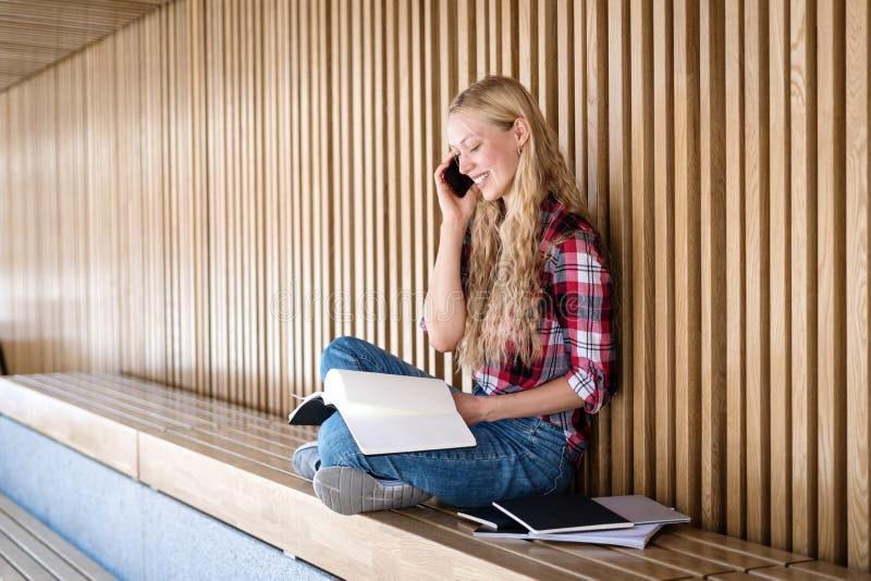 Side view of smiling adult student girl talking on modern smartphone, reading notes on copybooks with homework project, sitting on wooden bench near campus after class. Side view of smiling adult student girl talking on modern smartphone, reading notes on copybooks with homework project, sitting on wooden bench near campus after class